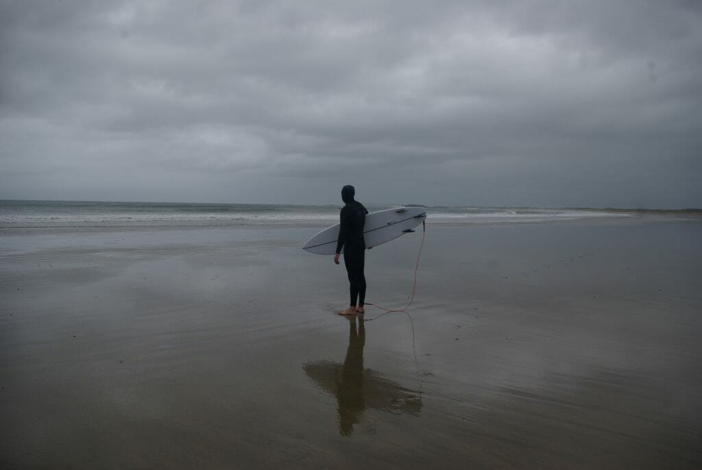 Surfen in Irland: Ein Surfer mit dickem Neo steht am Strand und spiegelt sich im nassen Sand.